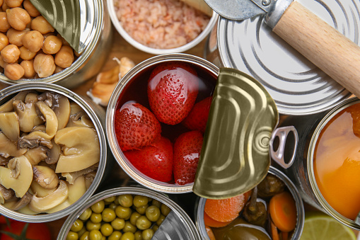 Open tin cans with different preserved products on wooden board, flat lay