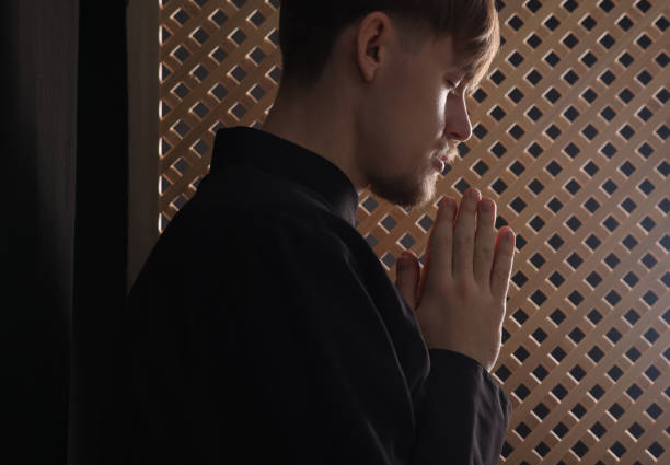 prêtre catholique priant près d’une fenêtre en bois dans une cabine confessionnelle - confession booth church forgiveness wood photos et images de collection