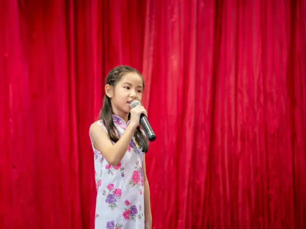 Photo of The Asian kid girl sing a song on stage at her school activity day, dress in Qipao style, red curtain background