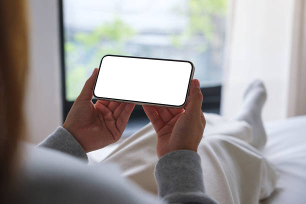 istock Mockup image of woman holding mobile phone with blank desktop white screen while sitting on a bed 1498039826