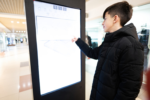 Teenager boy customer standing near electronic information desk looking for boutique or shop location in the big mall