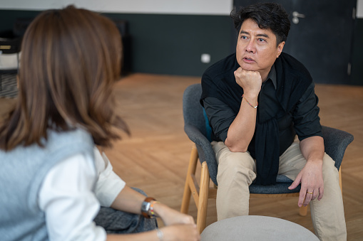 An Asian man sitting on cozy sofa and discussing faced problem with highly professional psychologist during therapy session at cozy office.