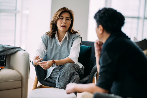 An Asian female professional psychologist and a mature man are sitting on a cozy sofa, discussing the problems he is facing during a therapy session at a comfortable office.
