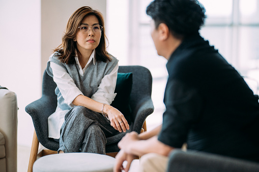 An Asian female professional psychologist and a mature man are sitting on a cozy sofa, discussing the problems he is facing during a therapy session at a comfortable office.