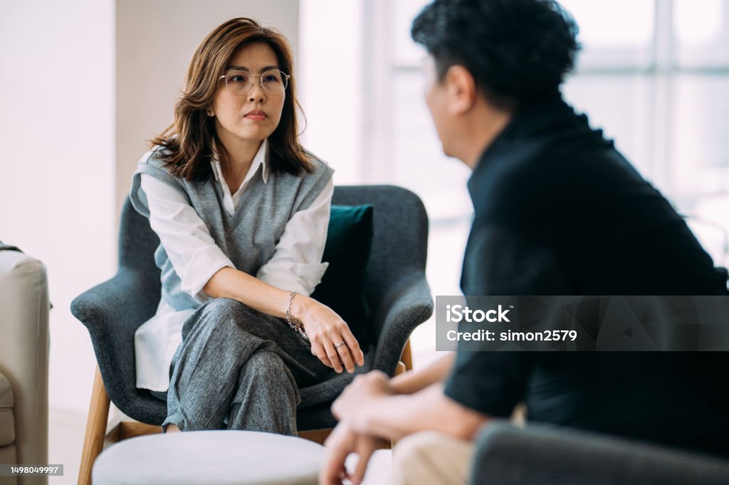Navigating Challenges: A Therapeutic Dialogue between an Asian Female Psychologist and a Mature Man - Consulting Patient at Modern Office An Asian female professional psychologist and a mature man are sitting on a cozy sofa, discussing the problems he is facing during a therapy session at a comfortable office. Mental Health Professional Stock Photo