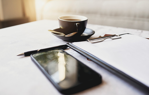 On a desk are a cup of coffee with a saucer and a spoon, a pen, a mobile phone, and a clipboard. Stock photo