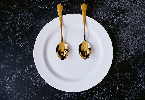 Top view of a white plate with two gold spoons shot on a charcoal background with copy space, stock photo