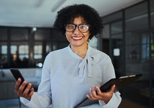 A businesswoman wearing eyeglasses stands holding her mobile phone and a file as she looks into the camera. Stock photo