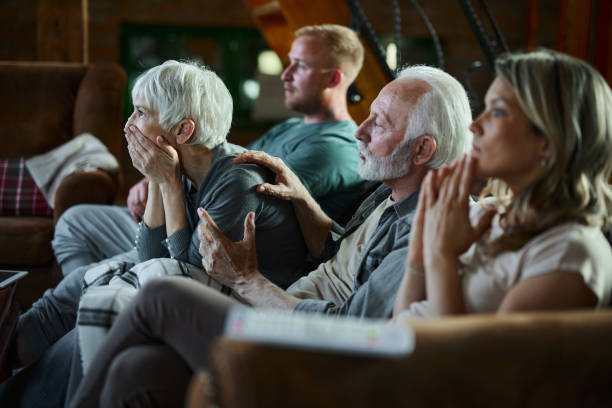 Watching a suspenseful movie at home! Senior parents and their adult children watching a movie in disbelief at home. Focus is on senior woman. suspenseful stock pictures, royalty-free photos & images