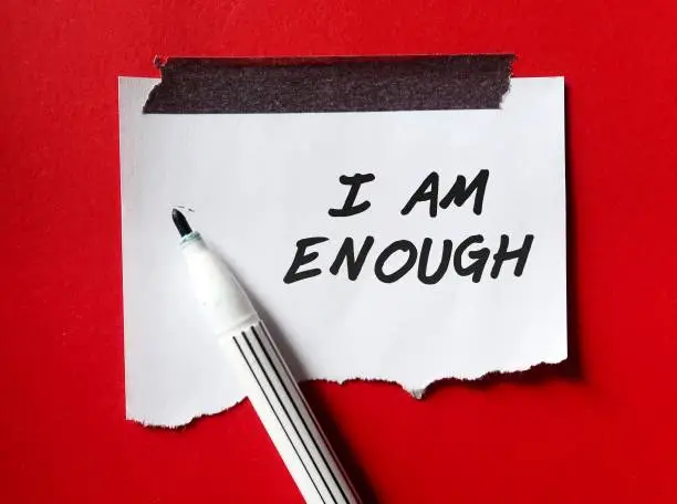 Stick note on red background with pen writing text I AM ENOUGH, Positive mantra affirmation to boost self acceptance, Knowing that you are worthy, valid, acceptable, loved. Embrace all flaws and imperfection
