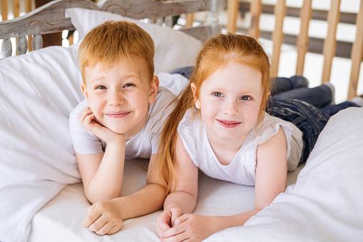 Red-haired boy and girl lie on a bed with white linens. Brother and sister are smiling and looking at the camera. Children portrait