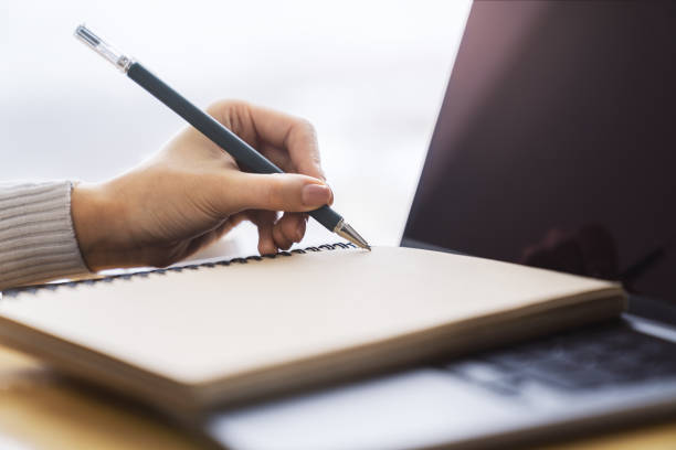istock Focused view of a woman's hand as she writes in a notepad on a modern laptop, background gently blurred 1498082832