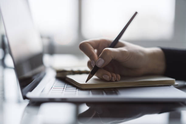 istock The image captures a woman's hand in close view as it writes in a notepad on a chic laptop, background out of focus 1498083735