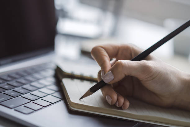 istock Tight focus on a woman's hand as she writes in a notepad on a contemporary laptop, background subtly blurred 1498083802
