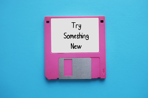 Pink floppy disk, diskette on blue background with text written TRY SOMETHING NEW, to encourage midlife unhappy workers or burnout employees to find inspiration in new pathways to career success