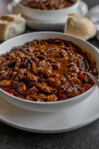 Delicious homemade high protein bean stew with kidney beans, baked beans, chicken meat and vegetables. Served ready to eat in a bowl with spoon on kitchen table with bread.