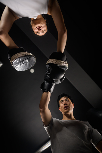Directly underneath male and female Chinese boxers hook and jab punching cross punches to pads in a boxing ring in Cape Town, South Africa.  The handsome Asian male is determined exercising hitting the hook & jab pads held by the young attractive Asian female in gym clothes.