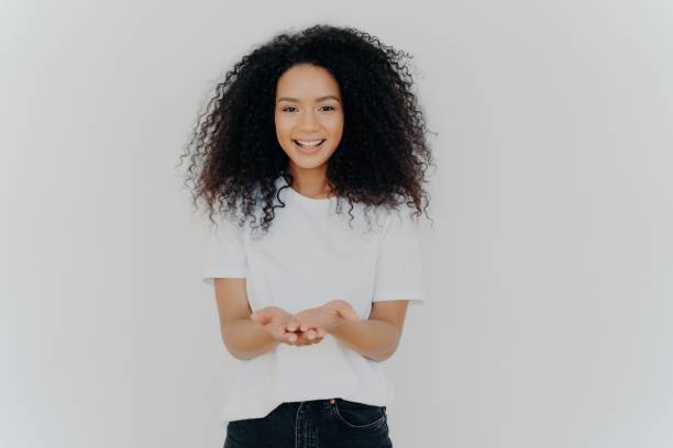 mujer alegre con las manos extendidas, pidiendo, sonriendo cálidamente, cabello encrespado, vistiendo camiseta y jeans, aislado en blanco. concepto de caridad. - frizzy 20s adult african descent fotografías e imágenes de stock