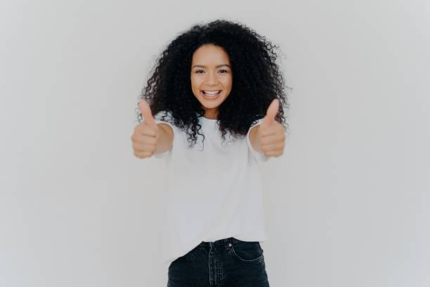 alegre mujer de cabello afro da el pulgar hacia arriba, aprueba con una sonrisa, en el interior, aislada sobre fondo blanco. - confidence toothy smile thumbs up ok sign fotografías e imágenes de stock