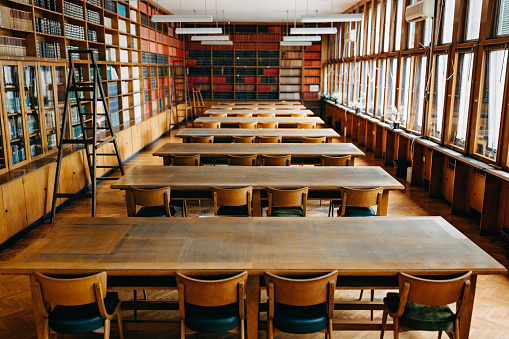 A photo captures the university library, showcasing its abundant collection of books neatly arranged on wooden bookshelves. Within the library, chairs and tables are placed in the center, surrounded by bookshelves on the sides.