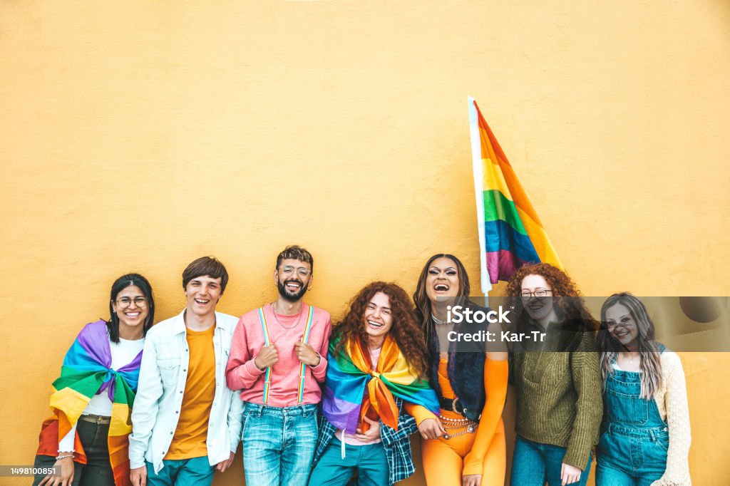 Diverse group of young people celebrating gay pride festival day - Lgbt community concept with guys and girls hugging together outdoors - Multiracial cheerful friends standing on a yellow background LGBTQIA Pride Event Stock Photo