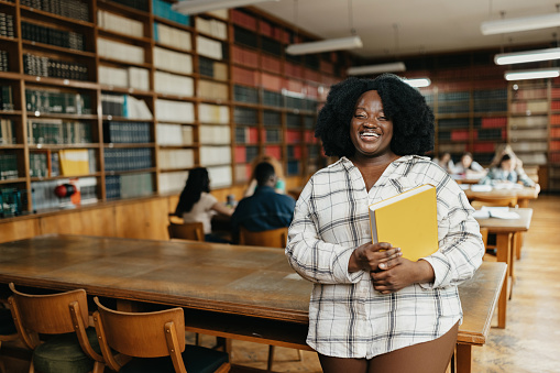Young african American female student holding book in the library while looking at camera