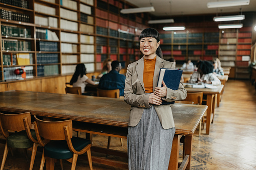 Mid adult Japanese ethnicity woman holding hard cover text book in the library while unrecognisable students are in the background