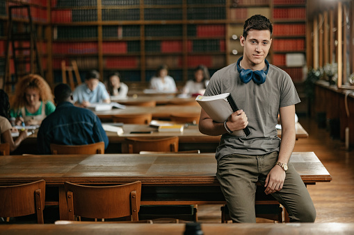 Portrait of the male student with headphones and text book in the public library while other students are in the background