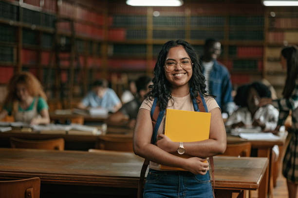 Beautiful Indian female student portrait while looking at camera Portrait of the Indian student holding a yellow book in the library education students stock pictures, royalty-free photos & images