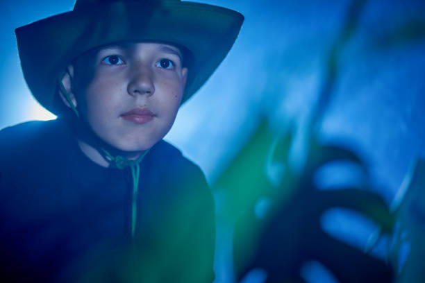 Cute preteen boy is looking away. Boy dressed in casual clothing and stetson hat, studio shooting in dark, shadows from house plants leaves
