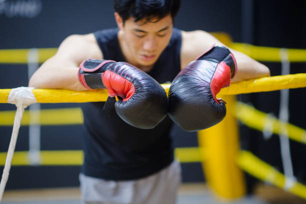 A Male Asian boxer is lying with arms resting on ropes looking tired want to give up but still has the boxing gloves on to give it a fight.