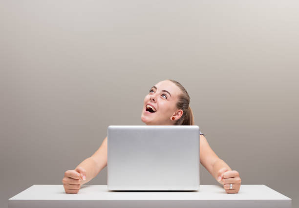 A beautiful young woman sits at a table with her laptop, her face expresses excessive joy and satisfaction, turned upwards to the copyspace. Computer, internet, positivity. Student