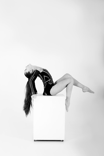 A beautiful girl gymnast in a black leotard does tricks on a white cube on a white background. White and black picture. Front view