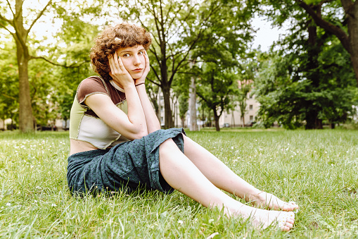 Portrait attractive happy smiling teenager girl, with curly brown hair, brown eyes, sitting on grass in park, barefoot, wearing shorts and T-shirt, summer season, sunny weather