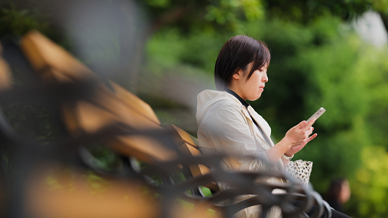 A beautiful woman is sitting at a bench and using her smart phone in city in a public park.