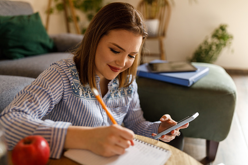 Portrait of diligent young female student sitting on the living room floor, at the coffee table, using smart phone while writing research and studying about nutrition.