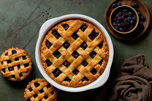 Whole baked mixed berry crust pie or tart with lattice, mini tarts. Homemade, fresh berries. Green table surface, directly above.