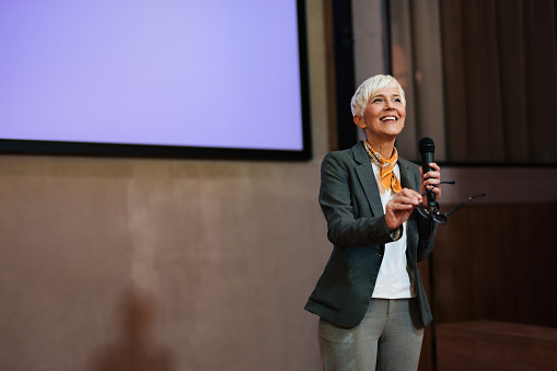 Smiling, ambitious senior businesswoman standing in front of the projection screen, giving a speech.