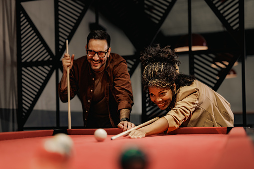 Cheerful African businesswoman playing billiard with a male colleague, having a great time after work.