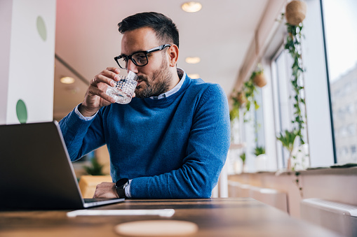 Serious male boss reading something on his laptop, drinking water, and sitting at the office.