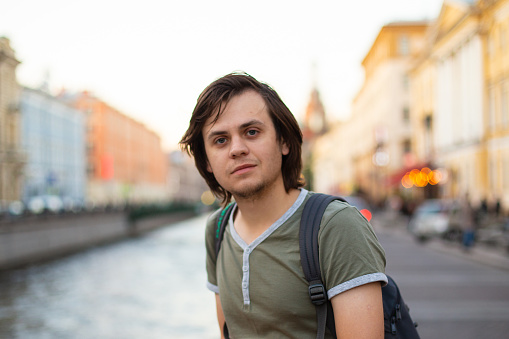 Young male man tourist with medium length hair in old city, river channel. European caucasian white man, looking at camera student, musician, violinist, painter, artist. Lifestyle portrait