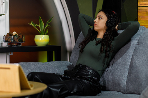Full length shot of cool teenage girl lounging on the sofa, hands behind head, enjoying listening to music via headphones. She is looking away, daydreaming.