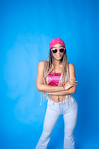 A Latin American woman standing against a blue background, wearing a pink headscarf over her long braided hair, pink sunglasses and a short pink top