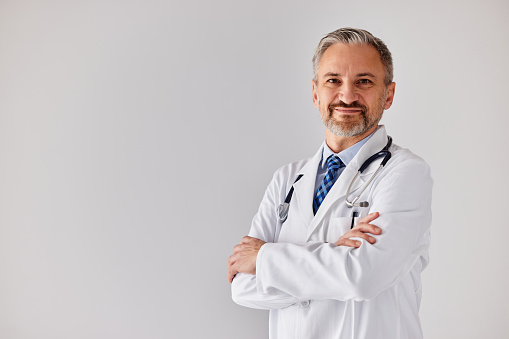 A smiling senior doctor in a white coat with a stethoscope, posing for the camera in front of the white wall.