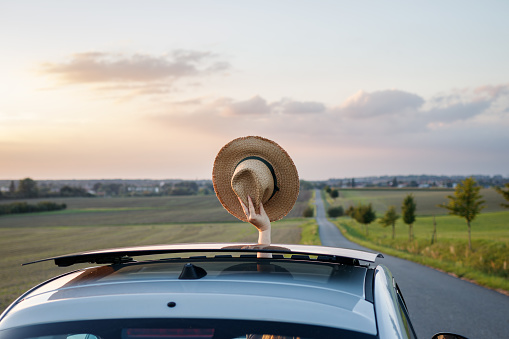 Travel by car on vacation. Woman waving with straw hat from car sun roof window during sunset. Summer road trip