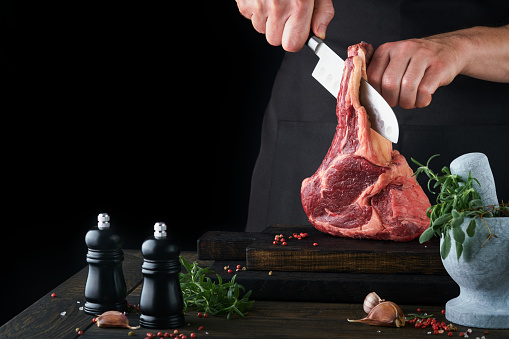 Chef cutting steak beef. Mans hands hold raw steak Tomahawk on rustic wooden cutting board on black background. Cooking, recipes and eating concept. Selective focus.