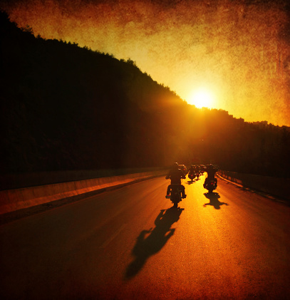 A large group of motorcycles are riding into the summer sunset.  The sun is ahead of them on top of a hill and the sun rays are shining onto the motorcycles and the road.  The sky is orange, as well as the road because if the sun reflection.