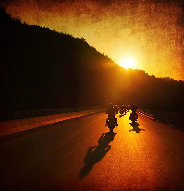 A large group of motorcycles are riding into the summer sunset.  The sun is ahead of them on top of a hill and the sun rays are shining onto the motorcycles and the road.  The sky is orange, as well as the road because if the sun reflection.