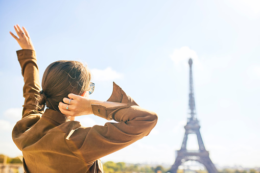 Female tourist raising hand and touching back of head, looking at blurred Eiffel Tower in distance