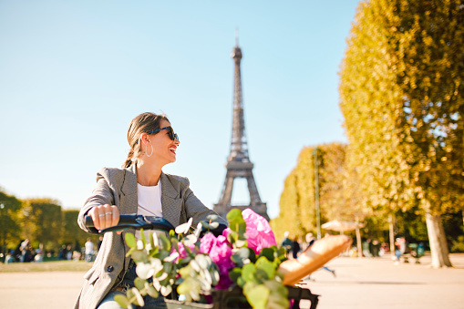 Female tourist riding electric bicycle, smiling and exploring park on sunny day, blurred view of Eiffel Tower in background